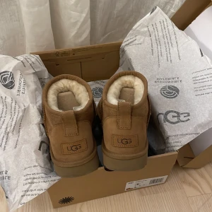 UGG boots i ljusbrun mocka med fårskinn - Mysiga UGG boots i modellen ultra mini och färgen: chestnut mocka med fluffigt vitt fårskinnsfoder. Skorna har rund tå, platt sula och klassisk UGG-logga baktill. Perfekta för kalla dagar och ger en chill vibe till din outfit. Obs! Står fast vid mitt pris💕