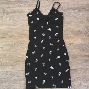 Svart fjärilsklänning från H&M Divided XS - Supersöt svart kortklänning med tunna axelband från H&M Divided. Klänningen har ett vitt fjärilsmönster, spetsdetaljer vid bysten och en liten rosett framtill. Den är figurnära och har en mjuk, stretchig känsla. Perfekt för dig som gillar unika detaljer och en feminin look.