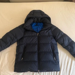 J.Lindeberg Vinterjacka - Snygg mörkblå dunjacka från J.Lindeberg med huva och två dragkedjeförsedda fickor. Jackan har quiltad design, svart yttertyg och blått innerfoder med tryckt logga. Perfekt för kalla dagar och har en modern, sportig look. Jackan är i helt nytt skick förutom att lite färg på dragkedjan har lossnat som ni ser på sista bilden. Denna modell säljs inte längre av J.Lindeberg. Nypris: 5499 kr