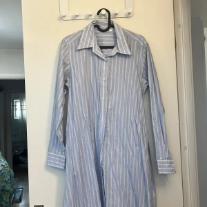 Blåvit randig skjortklänning - Snygg skjortklänning i blått och vitt med klassiska vertikala ränder. Långärmad modell med krage och knäppning hela vägen framtill. Tillverkad i mjuk bomull som känns skön mot huden. Perfekt för en chill och stilren look.