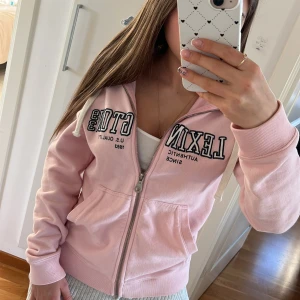 Lexington hoodie - Superfin ljusrosa hoodie från Lexington med dragkedja, huva och vita snören. Stor Lexington-logga broderad framtill. Mjuk bomullskvalitet och kängurufickor. 