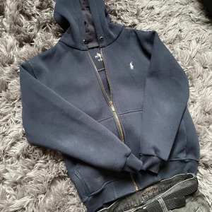 Ralph Lauren - Mörkblå hoodie från Polo Ralph Lauren med dragkedja och huva. Tröjan har broderad logga på bröstet, mjukt fleecefoder och ribbade muddar. Perfekt för kyliga dagar och enkel att matcha med jeans.