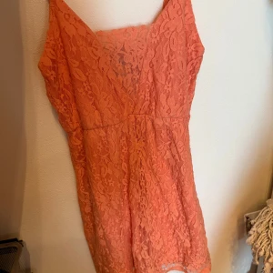 Korallfärgad spetsklänning - Säljer en superfin korallfärgad kort klänning med v-ringning och smala axelband. Klänningen är täckt av blommig spets och har en feminin och somrig vibe. Perfekt för dig som vill sticka ut lite extra! Materialet känns lätt och luftigt.