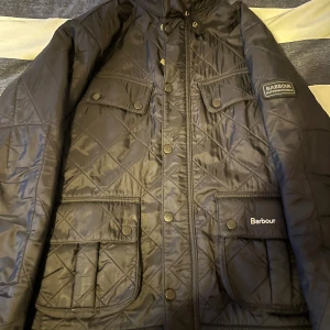 Svart quiltad jacka från Barbour  - Jackan är XL i barnstorlek det är därför det står XL skulle säga att den är ca XS annars .Quiltad svart jacka från Barbour International i storlek XL. Jackan har flera fickor med tryckknappar, dragkedja och Barbour-logga på bröstet. Klassisk look med rutmönster och hög krage. Perfekt för dig som gillar stilren och tidlös design.