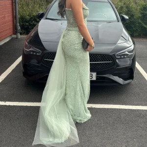 Ljusgrön glittrig balklänning - Magisk ljusgrön långklänning med glittriga detaljer och släp. Klänningen är axelbandslös med figurnära passform och har en snygg v-ringning framtill. Materialet är glittrigt och skimrande, perfekt för bal eller fest. Tyllsläp ger extra wow-effekt. 
