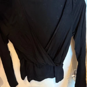 Svart omlottopp med lång ärm - Svart omlottopp med lång ärm och markerad midja. Toppen har en djup v-ringning framtill och är tillverkad i ett mjukt, stretchigt material som känns skönt mot huden. Perfekt för dig som gillar en enkel men snygg look.