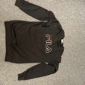 Svart sweatshirt från FILA - Svart sweatshirt från FILA med stort logotryck i vitt och rött på bröstet. Tröjan har rund halsringning, långa ärmar och ribbade muddar vid ärmslut och nederkant. Perfekt för en sportig och avslappnad stil.