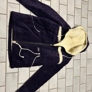 Mörklila jacka med teddyfoder - Snygg mörklila jacka från Sting med mysigt teddyfoder i hela luvan och insidan. Jackan har dragkedja, snörning i midjan och två fickor framtill. Perfekt för kalla dagar när du vill vara både varm och stilren.