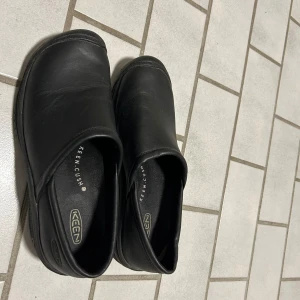 Svarta slip-on skor från Keen - Svarta slip-on skor från Keen med rund tå och platt sula. Skorna är tillverkade i skinn och har en enkel, stilren design med diskret logga på innersulan. Perfekta för dig som gillar minimalistisk stil och komfort.