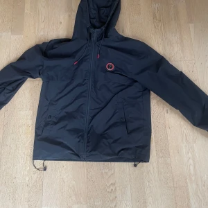 Trapstar windbreaker jacka - Svart vindjacka från Trapstar med huva och dragkedja. Jackan har röd broderad Trapstar-logga på ryggen och en rund logga på bröstet. Justerbar dragsko nertill och vid huvan, samt två fickor framtill. Perfekt för dig som vill ha streetwear-vibe.