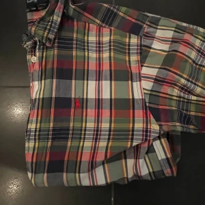 Rutig skjorta från Ralph Lauren - Färgglad rutig skjorta från Ralph Lauren med klassisk krage och knäppning framtill. Skjortan har långa ärmar och är tillverkad i mjuk bomull. Färgerna går i blått, grönt, rött, gult och vitt med den ikoniska röda loggan broderad på bröstet.