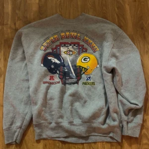 Vintage sweatshirt  - Säljer en grå sweatshirt från Pro Player med stort Super Bowl XXXII-tryck framtill, featuring Broncos och Packers. Tröjan är i klassisk loose fit med rund hals och långa ärmar. Materialet är en mix av bomull och polyester, perfekt för dig som gillar sportig vintage.