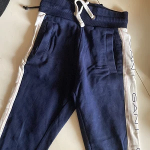 Marinblå mjukisbyxor från Gant - Mörkblå mjukisbyxor från Gant med vita sidor och GANT-logga längs benen. Byxorna har snörning i midjan och ribbade muddar. Perfekta för chill dagar eller sport. Tillverkade i mjukt bomullsmaterial för extra komfort.