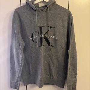 Grå hoodie från Calvin Klein Jeans - Säljer en grå hoodie från Calvin Klein Jeans med stor CK-logga på bröstet. Tröjan har huva med snörning och ribbade muddar vid ärmslut och nederkant. Perfekt för en chill och stilren look.