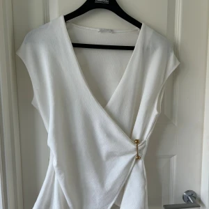 Vit omlottopp med gulddetalj - Säljer en stilren vit omlottopp med djup v-ringning och ärmlös design. Toppen har en snygg guldfärgad detalj i sidan som håller omlottstilen på plats. Toppen sitter rätt tjugu och känns lite stickad 