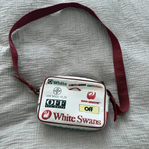 Vit axelväska Off-White White Swans - Säljer en vit axelväska från Off-White med röd axelrem och flera tryck. Inget slitage på väskan och dragkedjan funkar som ny. Köptes 2020 från farfetch för 8700kr. Väskan är i väldigt bra skick då den knappt har använts.