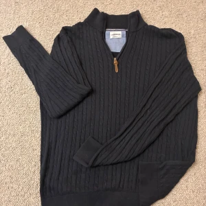 Kabelstickad halfzip tröja från Lindbergh - Säljer en mörkblå kabelstickad tröja från J.Lindbergh med relaxed fit. Tröjan har halv dragkedja med snyggt läderdetalj, ribbade muddar och hög krage. Perfekt för dig som gillar klassisk och stilren look.