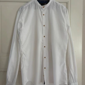 Vit henley-skjorta från Zara - Vit henley-skjorta från Zara i bomull med snygga träfärgade knappar och ståkrage. Långärmad modell med coola detaljer på insidan av manschetterna och kragen i blått tyg med mönster. Perfekt för en clean och stilren look.