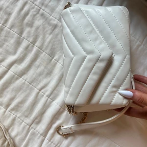 Vit quiltad axelväska med guldkedja - Säljer en stilren vit axelväska med quiltad design och snygg guldfärgad kedja. Väskan har ett huvudfack med dragkedja inuti och magnetlås. Perfekt storlek för det viktigaste och passar till många outfits.