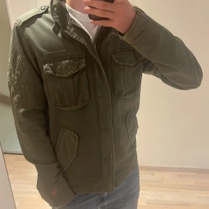 Vintage grön jacka  - Säljer en olivgrön parkajacka med fyra stora fickor framtill och coola detaljer på ärmarna. Jackan har hög krage, axelklaffar och stängs med dragkedja och knappar. Perfekt för dig som gillar en avslappnad och stilren look. Storleken är L men passar som en M