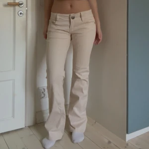 Beiga bootcut byxor från American Flavour - Säljer ett par beiga bootcut-byxor från American Flavour med diskret randigt mönster. Byxorna har låg midja, klassiska fickor och är tillverkade i bomullsmix med lite stretch. Perfekta för dig som gillar retrovibbar och vill ha en snygg siluett. Jag är 170 cm lång och innerbenslängden är 81 cm. Midjemåttet rakt över är 40 cm 