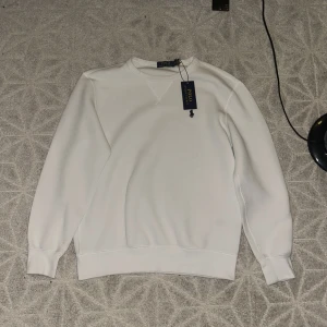Vit sweatshirt från Polo Ralph Lauren - Säljer en stilren vit sweatshirt från Polo Ralph Lauren med klassisk rund hals och broderad logga på bröstet. Tröjan har långa ärmar, ribbade muddar och är tillverkad i mjuk bomullsmix. Perfekt för en clean och avslappnad look.