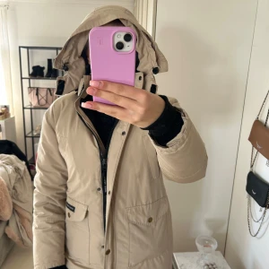 Beige parkas från Canada Goose - Snygg beige parkas från Canada Goose med huva och klassisk logga på ärmen. Jackan har stora fickor framtill, dragkedja och knappar. Perfekt för kalla dagar och har en clean, stilren look som funkar till allt.