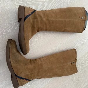Stövlar från Tommy Hilfiger - Snygga bruna stövlar i mocka från Tommy Hilfiger med dragkedja på insidan och en diskret blå och röd detalj vid hälen. Platt sula och klassisk rund tå gör dem perfekta till många outfits. Stövlarna har en stilren look och är lätta att matcha. Molly 10B.