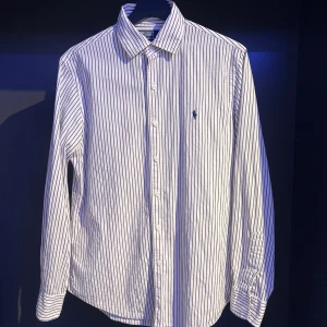Randig skjorta från Polo Ralph Lauren - Säljer en klassisk vit skjorta med smala svarta ränder från Polo Ralph Lauren. Skjortan har lång ärm, knappar framtill och den ikoniska broderade loggan på bröstet. Modellen är Custom Fit och passar perfekt till dig som gillar stilrena plagg. Skick 9/10 och den har knappt använts. Har man några frågor eller funderingar är det bara att höra av sig 👍