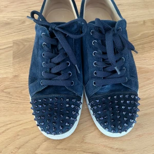 Mörkblå mocka sneakers med nitar - Säljer ett par mörkblå sneakers i mocka med coola svarta nitar på tån. Skorna har snörning och vit sula, vilket ger en snygg kontrast. Perfekta för dig som vill sticka ut med en edgy vibe och gillar detaljer som verkligen syns.