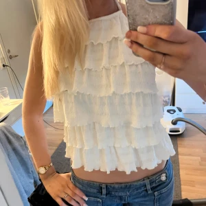 Vit volangig ärmlös linnetopp - Säljer en söt vit linnetopp med flera lager volanger och hög hals. Toppen är ärmlös och croppad, perfekt för varma dagar. Materialet känns lätt och luftigt, vilket gör den superbekväm att bära. Passar snyggt till jeans eller kjol.