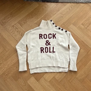 Vit polotröja med text och knappar - Vit stickad polotröja med bred ribbad krage och coola knappar längs axeln. På framsidan står det 'ROCK & ROLL' i vinröda bokstäver. Tröjan har långa ärmar och extra breda muddar för en trendig look.