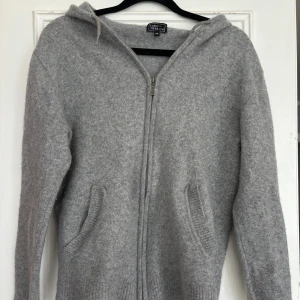 Grå hoodie i kashmir från CASHMERE.CH - Mysig grå hoodie i kashmir från CASHMERE.CH med dragkedja framtill, huva och två fickor. Tröjan har hål på armbågarna