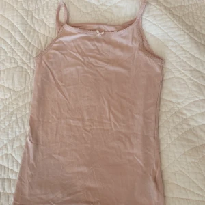 Ljusrosa linne med rosett från H&M - Supersött ljusrosa linne från H&M med smala axelband och en liten rosett framtill. Toppen har spetsdetaljer längs kanten och är gjord i mjuk bomull, perfekt för varma dagar eller som layering-piece.