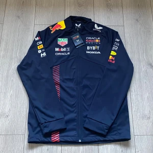 Red Bull Racing jacka från Castore  - Mörkblå Red Bull Racing jacka från Castore i storlek M. Jackan har tryckta loggor från Red Bull, Mobil 1, Oracle och fler sponsorer, samt röda detaljer på framsidan. Långärmad med dragkedja och sportig look, perfekt för motorsportfans.