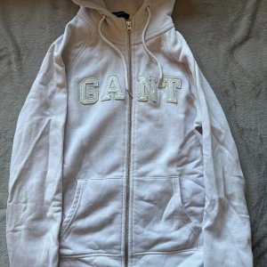 Ljusrosa hoodie från Gant - Säljer en ljusrosa hoodie från Gant med dragkedja och broderad logga framtill. Tröjan har huva med snörning och två fickor framtill. Perfekt för en chill och avslappnad stil.