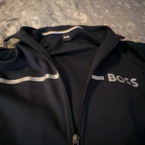 Svart ziptröja från BOSS med logga - Svart långärmad ziptröja från BOSS med vit vågig detalj och stor BOSS-logga på bröstet. Perfekt för en clean och sportig look.