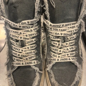 Zadig & Voltaire grå high-top sneakers - Unika high-top sneakers från Zadig & Voltaire i grått canvasmaterial med slitna, fransiga kanter och vita snören med logotyp. Sidan har en silverfärgad dragkedja och sulan är vit med vintage-look. Perfekta för dig som gillar edgy streetstyle.