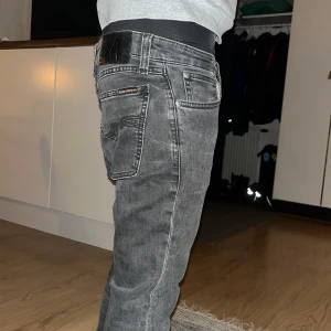 Svarta jeans från Nudie Jeans Co - Säljer ett par svarta jeans från Nudie Jeans Co med snyggt tvättad look och klassiska bakfickor med broderad detalj. Jeansen har normal passform och raka ben, tillverkade i mjuk bomullsdenim. Perfekta för dig som gillar stilrena och bekväma jeans.