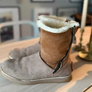 Canada Snow mockaboots med fårfoder - Mysiga varma boots från Canada Snow i ljusbrun äkta mocka med vitt fårfoder. Skorna har dragkedja på sidan och en platt sula med mönster av lönnlöv för extra grepp. Perfekta för kalla dagar och har en klassisk, rund tå och robust känsla. Storlek 40, upplever dem som normala i storlek.  Mockan är lite "ruggig" på ytan och mönstret under sulan är lite nedslitet bak. Inga andra anmärkningar.