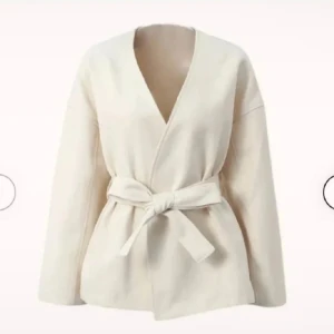 Beige omlottjacka med bälte - Stilren beige omlottjacka med knytbälte i midjan och v-ringad hals. Jackan har lång ärm och en avslappnad passform, perfekt för lager-på-lager. Tillverkad i mjukt material som ger en clean och modern look.