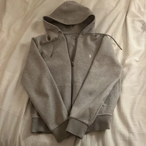 Grå hoodie från Polo Ralph Lauren - Snygg grå hoodie från Polo Ralph Lauren med dragkedja och klassisk broderad logga på bröstet. Tröjan har huva med snörning, långa ärmar och ribbade muddar. Perfekt för chill dagar och enkel att matcha med jeans eller joggers.| Tröjan är använd typ 3 gånger, säljes då den är för liten| kan gå ner i pris☺️