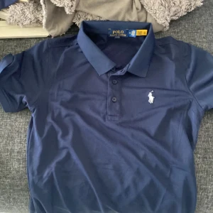 Marinblå pikétröja från Ralph Lauren - Snygg marinblå pikétröja från Polo Ralph Lauren med klassisk krage och knäppning framtill. Tröjan har korta ärmar och den ikoniska vita broderade loggan på bröstet. Perfekt för en stilren och sportig look.