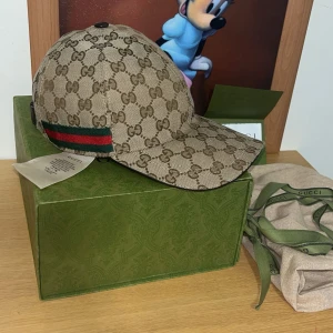Gucci keps med GG-mönster - Säljer en beige keps från Gucci med klassiskt GG-monogram över hela kepsen och grönt/rött band på sidan. Kepsen har böjd skärm och är tillverkad i canvas med detaljer i bomull. Perfekt för dig som vill sticka ut med en lyxig accessoar.