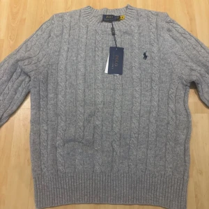 Grå kabelstickad tröja Polo Ralph Lauren - Kabelstickad grå tröja från Polo Ralph Lauren med rund halsringning och broderad marinblå logga på bröstet. Tröjan har ribbade muddar och är tillverkad i mjukt material som passar perfekt för kyligare dagar.