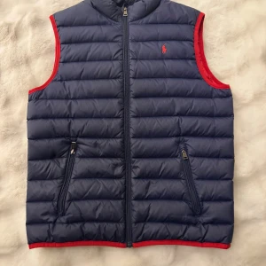 Blå dunväst från Polo Ralph Lauren - Mörkblå quiltad dunväst från Polo Ralph Lauren med röda detaljer och dragkedja framtill. Västen har två fickor med dragkedja och röd broderad logga på bröstet. Insidan är helröd och västen har en sportig, klassisk look.