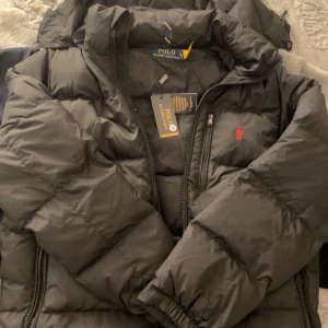 Svart pufferjacka från Polo Ralph Lauren - Svart pufferjacka från Polo Ralph Lauren med röd broderad logga på bröstet. Jackan har hög krage, dragkedja framtill och sidofickor med dragkedja. Tillverkad i syntetmaterial och perfekt för kalla dagar. Klassisk och stilren design.