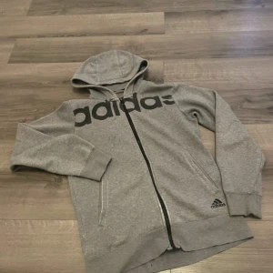Grå hoodie från Adidas med dragkedja - Säljer en grå hoodie från Adidas med svart logga över bröstet och mindre logga vid fickan. Tröjan har huva, långa ärmar, två fickor och hel dragkedja framtill. Perfekt för chill eller träning. Materialet är mjuk bomullsblandning.