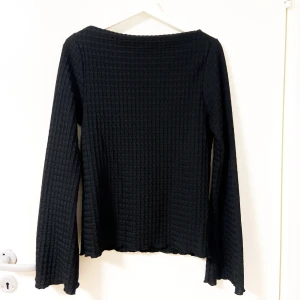 Svart långärmad topp från 157 - Svart långärmad topp från 157 med vågig struktur i tyget och lätt utsvängda ärmar. Toppen har en rak passform och är tillverkad i ett stretchigt material som känns bekvämt mot huden. Perfekt att styla med jeans eller kjol för en clean look.