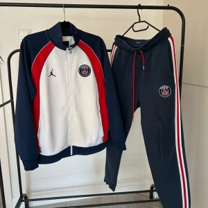 PSG x Jordan träningsset M - Säljer ett fett PSG x Jordan träningsset i storlek M. Setet består av en zip-jacka och matchande byxor i blått, vitt och rött med Paris Saint-Germain-logga och Jordan-logga. Byxorna har röda och vita ränder längs sidorna och dragsko i midjan. Perfekt för dig som älskar fotboll och streetstyle.
